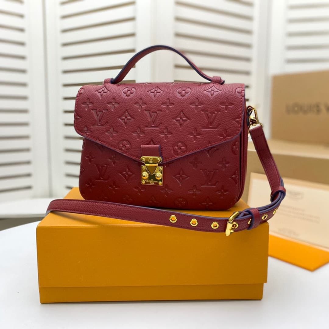 A Louis Vuitton monogram embossed leather handbag in burgundy red, displayed on top of a yellow-orange Louis Vuitton box with its matching shoulder strap laid across the front. The background shows white louvered shutters and a cardboard box.